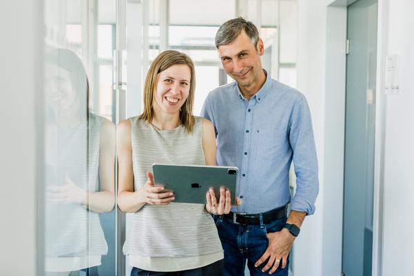 A female and a male consultant standing in a hallway with glass walls. The female is holding an iPad discussing client requests.